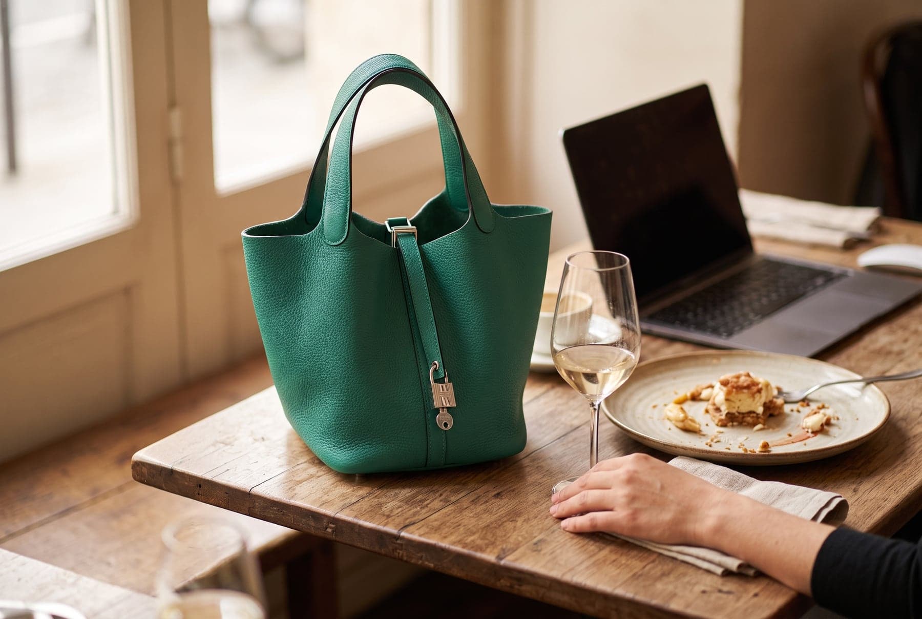 Green Hermès Picotin Lock MM bag with silver padlock on a rustic wooden café table beside a wine glass, dessert plate and laptop.
